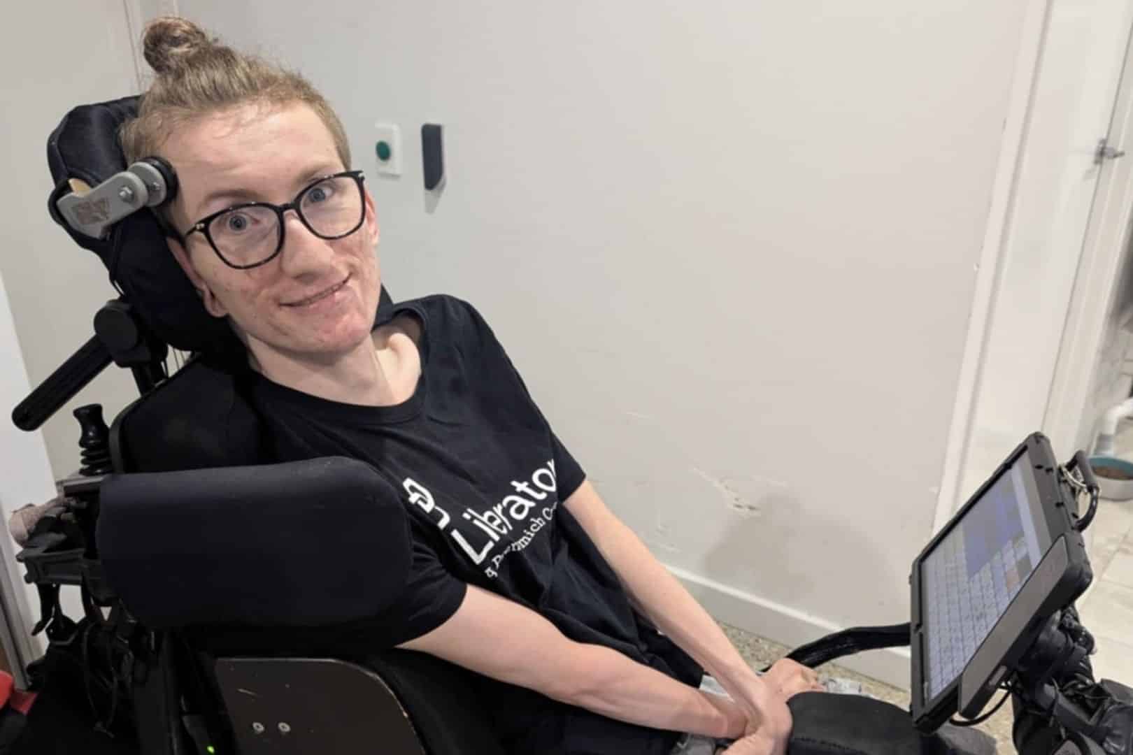 A Caucasian male with glasses sits in a mobility chair with an aac device and smiles for the camer