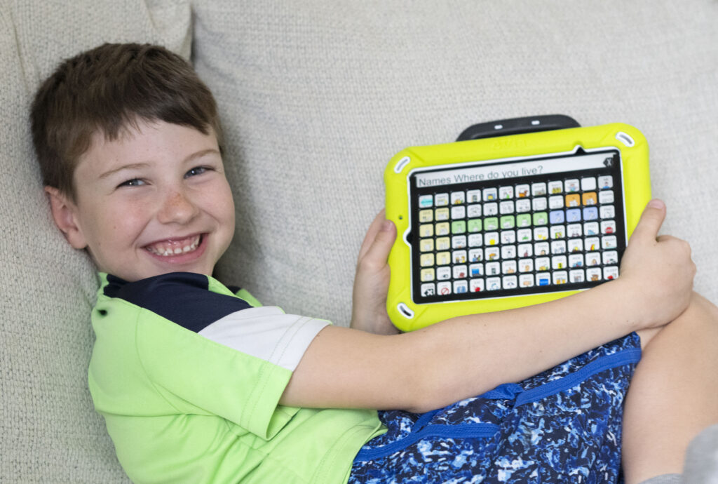 Young boy smiles and holds a yellow AAC device with Unity® app